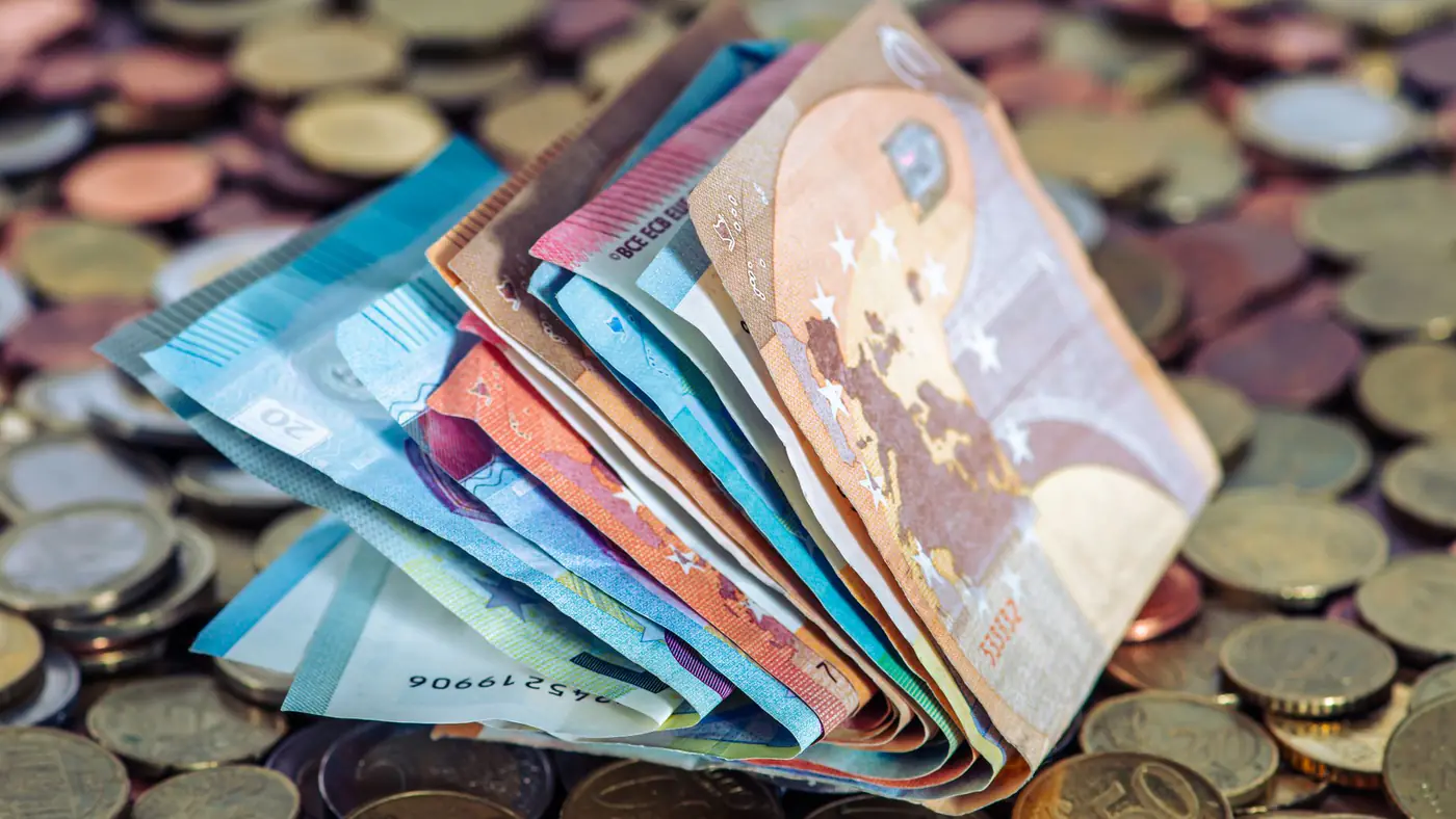 A stack of euro banknotes and scattered coins symbolize wealth and finance.