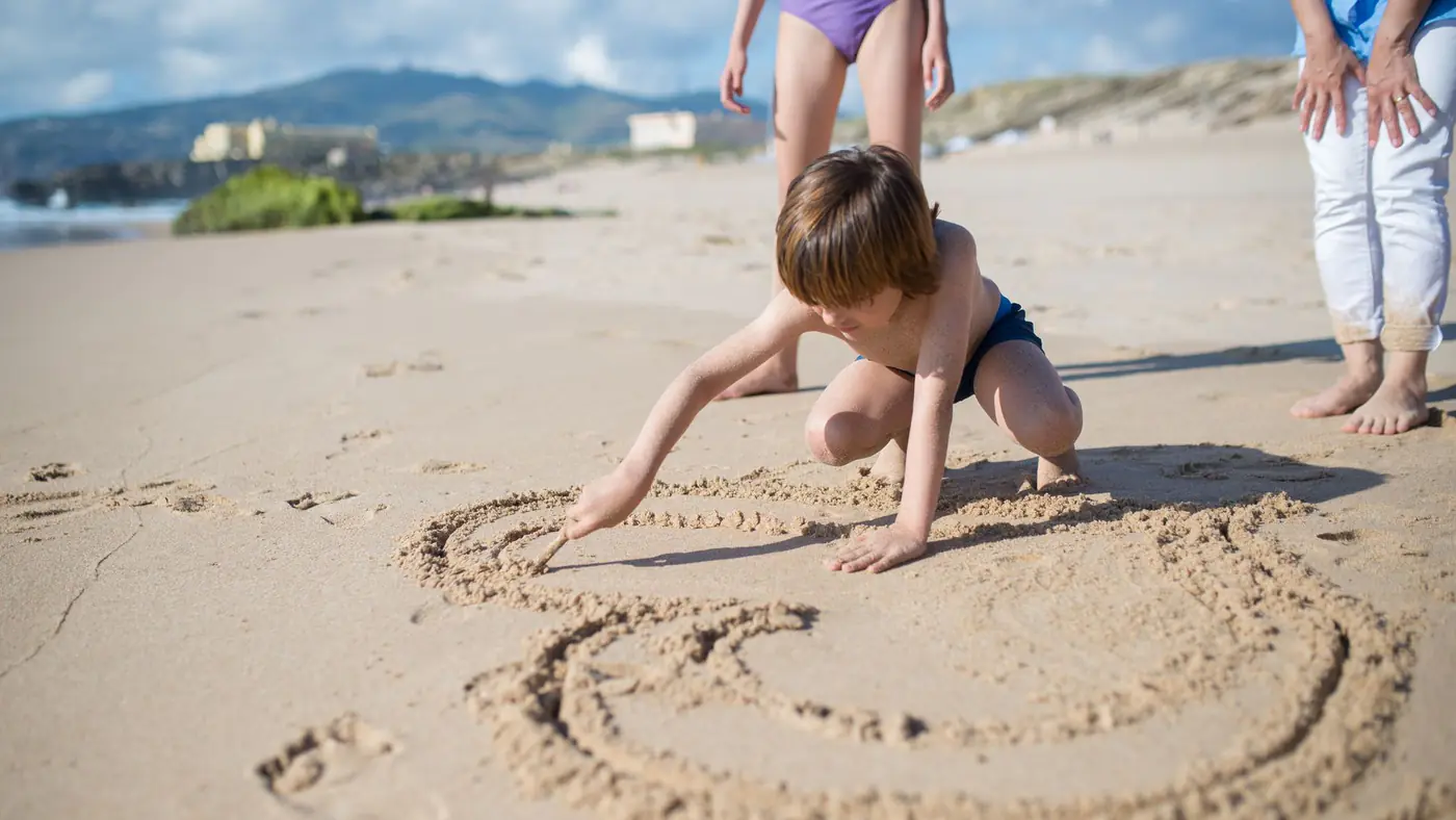 A child enjoys drawing in the sand on a sunny beach in Portugal, capturing a fun vacation moment.
