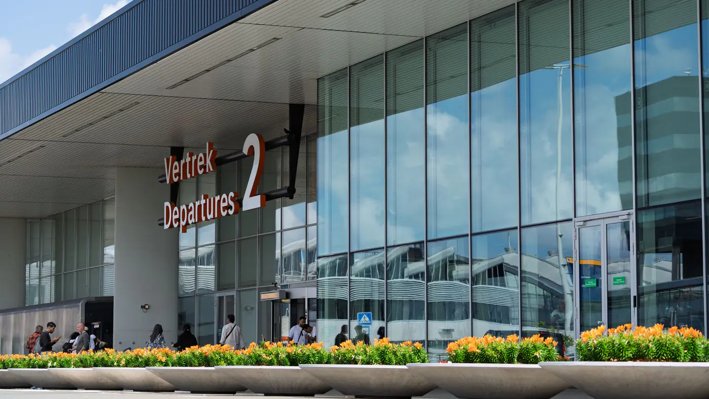 Glass facade of a bustling airport departure terminal with people outside and reflection of clouds.