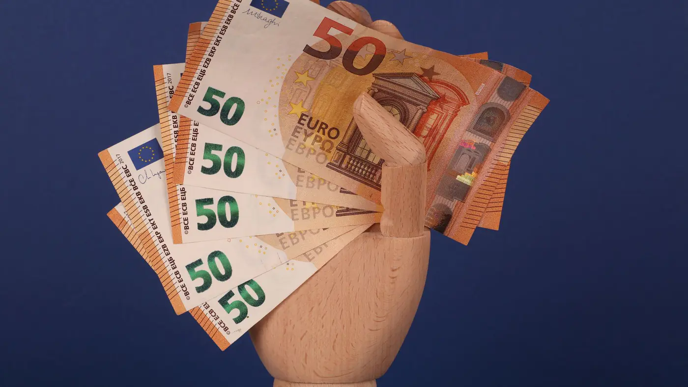 A wooden mannequin hand grasping fifty euro banknotes with a blue backdrop.