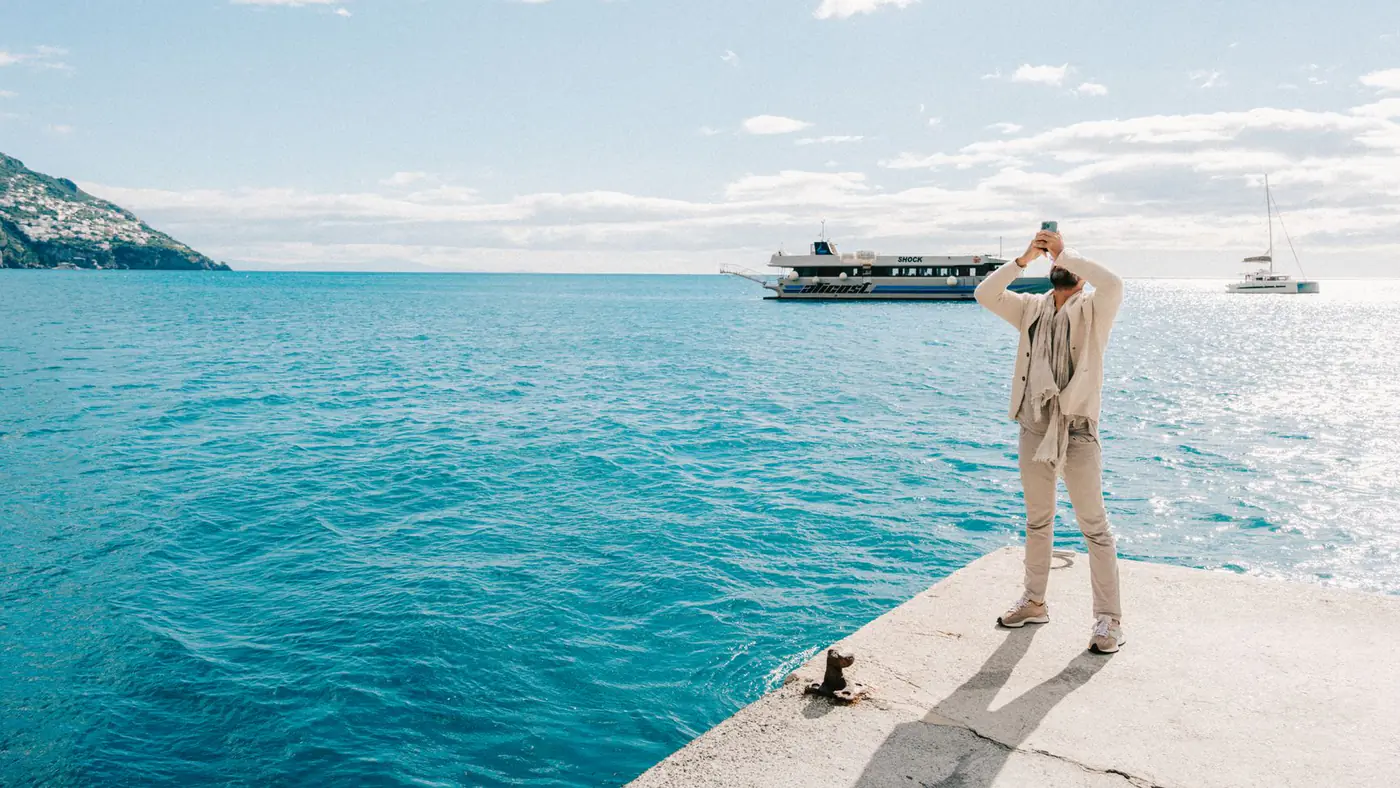 A man stands at a sunny seaside capturing the ocean view with ships in the distance.
