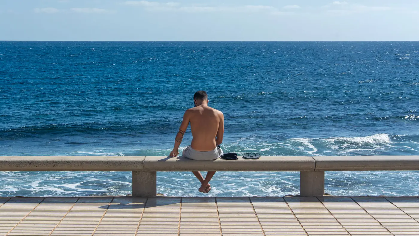 A shirtless man sitting on a bench, gazing at the ocean under a clear sky.
