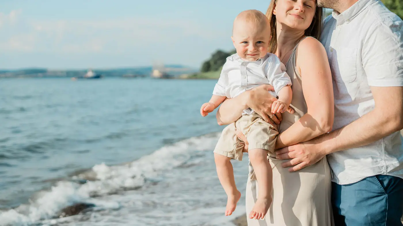A joyful family moment captured by the sea, with a couple holding their smiling baby on a sunny day.