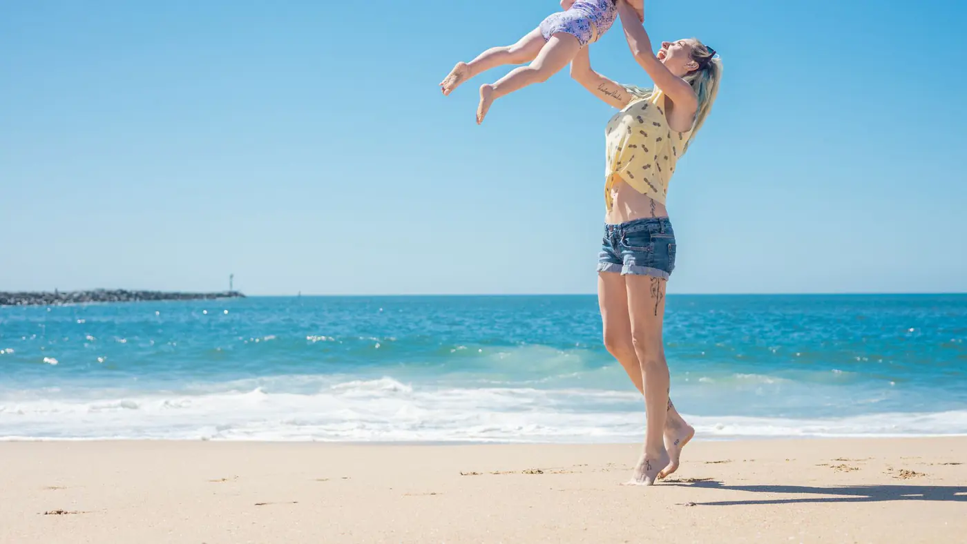 A mother joyfully lifts her daughter in the air at a sunny beach, capturing a moment of family fun.
