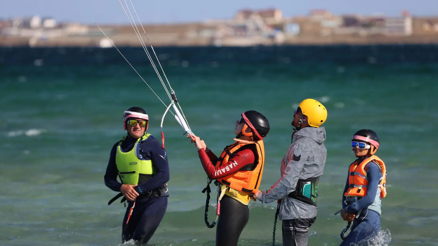 Adults and teens taking a kiteboarding lesson on a sunny beach, wearing safety gear and wetsuits.