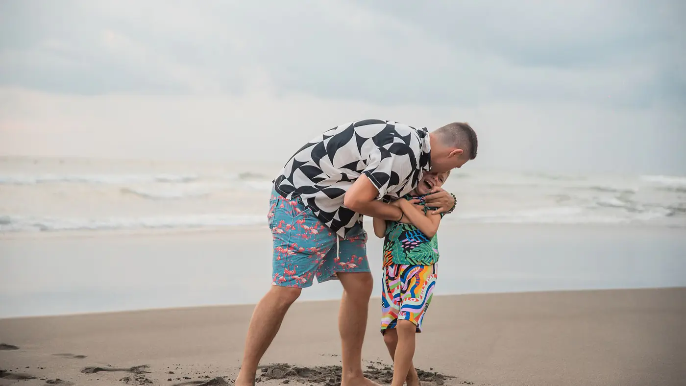 A joyful interaction between father and son playing on a sandy beach under a cloudy sky.