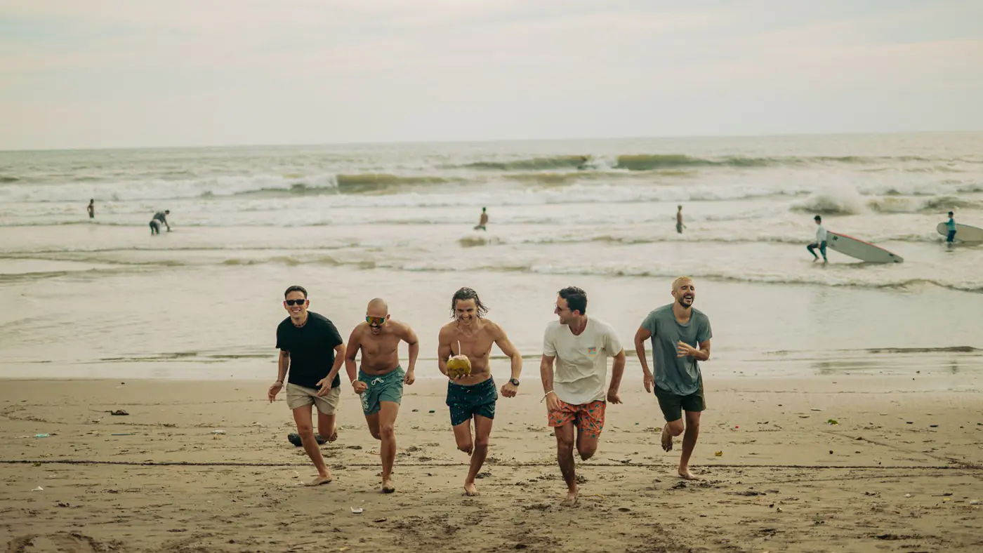 Five friends running on a sandy beach, enjoying a sunny day with surfers in the ocean.
