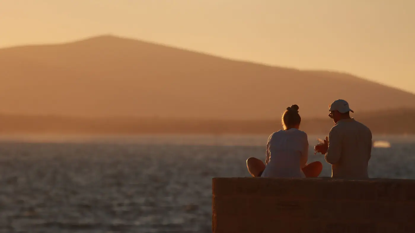 Couple enjoying a serene sunset by the shore in Alghero, Sardinia, Italy.