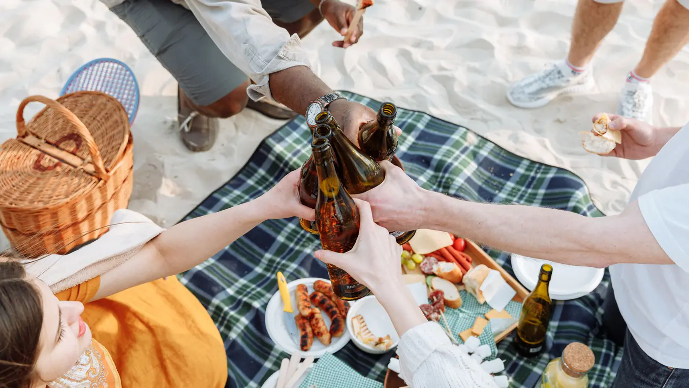 A joyful picnic on the beach with friends clinking bottles and enjoying food.