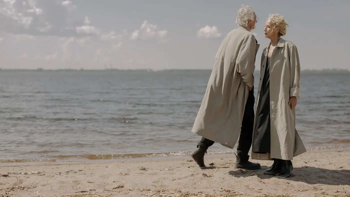 An elderly couple in long coats walks along a serene beach under a clear blue sky.