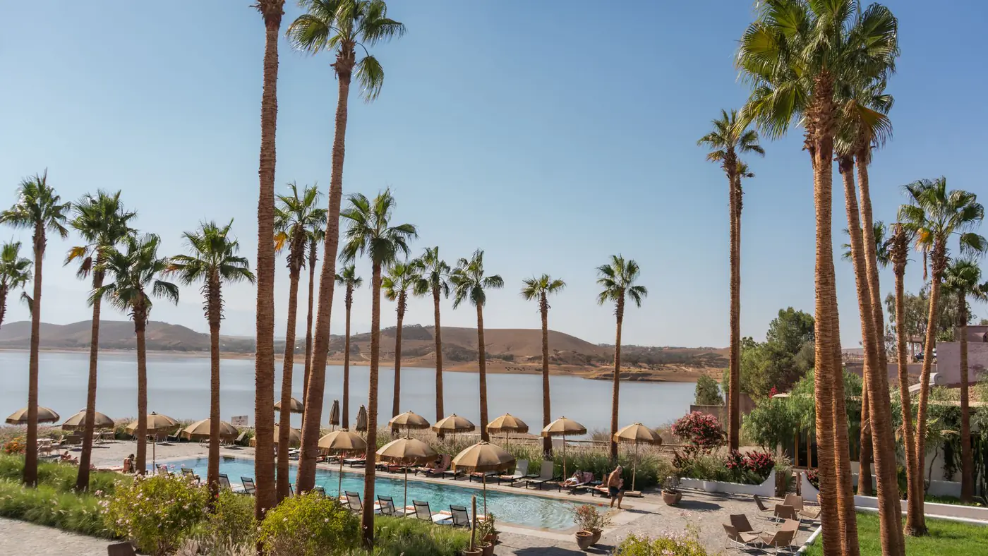 Tranquil resort setting featuring palm trees, a pool, and a stunning lake view under a clear blue sky.
